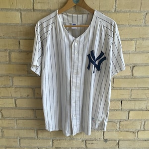 May include: White baseball jersey with navy blue pinstripes and a navy blue "NY" logo on the left chest. The jersey has short sleeves and a button-up front. It is hanging on a wooden hanger against a brick wall.