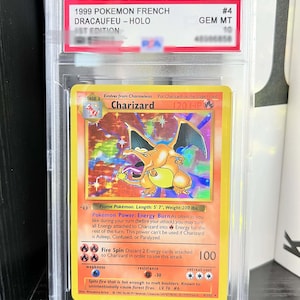 May include: A 1999 Pokemon Charizard trading card, graded GEM MT 10 by PSA, encased in a clear plastic slab. The card features a holographic illustration of the orange dragon-like creature, with text including Charizard, 120 HP, and Fire Spin.