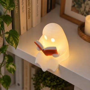 May include: A white, ghost-shaped lamp emitting a warm, soft glow from within. The ghost figure is seated on a ledge, holding an open book with orange covers. The lamp features two circular cutouts for eyes, allowing light to shine through.