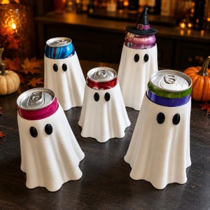 May include: A set of five white ghost-shaped beverage can covers, each featuring two black oval eyes. One cover includes a small black witch hat on top. The covers are designed to fit over standard metal drink cans, with one can displaying the text VIKING BERRY.