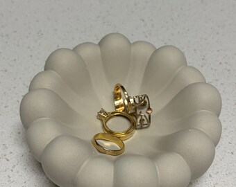 Puede incluir: Un cuenco de color blanco roto mate con borde festoneado que contiene tres anillos de oro. Uno de los anillos tiene una pequeña piedra transparente engastada, mientras que los otros dos son bandas de oro lisas y sencillas.
