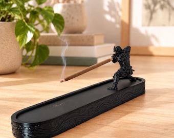 Puede incluir: Un quemador de incienso negro con una figura de samurái esculpida en una pose dinámica. La figura sostiene una varilla de incienso marrón que emite un fino rastro de humo. La base es de forma ovalada con intrincados relieves decorativos.
