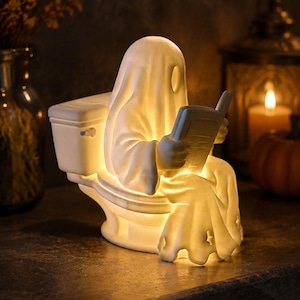 May include: A white ceramic ghost figurine sitting on a toilet while reading an open book. The ghost is illuminated from within, casting a warm glow through its translucent material. The figure features a draped sheet design and a simple oval face.