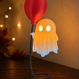 May include: A white, glowing ghost figurine holding a red balloon. The ghost is attached to a thin, dark, curved metal stand that rises from a circular base. The ghost features two oval-shaped cutouts for eyes that emit a warm, soft light.