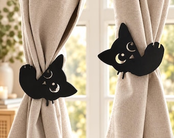 Puede incluir: Dos alzapaños de cortina de metal negro con forma de siluetas de gato. Cada figura de gato presenta detalles recortados en forma de luna creciente y estrella en la cara y sujeta un panel de cortina de color beige con textura de lino.