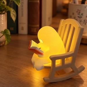 May include: A glowing, yellow, ghost-shaped lamp sitting in a small, light-colored rocking chair. The figure is holding an open book with orange covers and white pages, illuminated by the warm light emitted from the lamp's body.