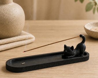 Puede incluir: Un porta-incienso negro de forma ovalada con una figura de gato esculpida en un extremo. La bandeja tiene un borde estriado y sostiene una varilla de incienso encendida que se extiende diagonalmente desde la cola del gato, soltando humo.