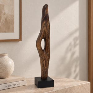 Può includere: Una scultura in legno marrone scuro, alta e allungata, con una forma organica liscia caratterizzata da un'apertura ovale centrale. La scultura è montata verticalmente su una base quadrata in nero opaco, che mette in risalto le venature naturali del legno.