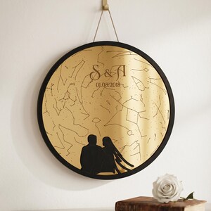 May include: A circular wall hanging featuring a gold reflective surface with a black star map constellation design. The center displays the text S & A and the date 01.08.2018. A black silhouette of two people sits at the bottom of the frame.
