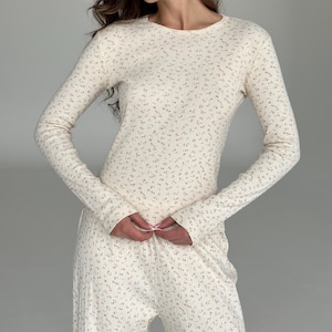 Può includere: Un completo composto da una maglia a maniche lunghe con girocollo e pantaloni abbinati, in tessuto a coste color bianco sporco. Il capo presenta un delicato motivo ripetuto di minuscoli fiori rosa e verdi su tutta la superficie.