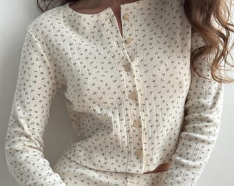 May include: A cream-colored long-sleeve button-down top featuring a delicate dark floral pattern and a ribbed texture. The garment has a round neckline and a vertical row of small, light-colored buttons running down the center front.