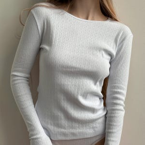 Peut inclure: Un haut blanc à manches longues présentant une texture côtelée verticale subtile avec un motif pointelle délicat. Le vêtement possède une encolure ronde et une silhouette ajustée, mettant en valeur un design monochrome épuré.