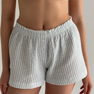 Puede incluir: Pantalones cortos a rayas azul claro y blanco con un acabado de tela texturizada y arrugada. Los pantalones cortos tienen una cintura elástica con volantes y una tapeta frontal adornada con tres botones blancos pequeños y redondos.