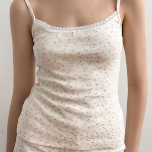 Puede incluir: Una camiseta de tirantes acanalada de color crema con un delicado estampado floral en rosa y verde. La prenda tiene tirantes finos, un escote redondo adornado con encaje blanco y un pequeño lazo central en la parte delantera del escote.