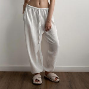 May include: White loose-fitting pants made of a textured, crinkled fabric. The pants feature an elasticized waistband with a matching white drawstring tie at the center front. The legs have a relaxed, wide-leg silhouette that tapers slightly at the hem.