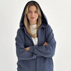 Può includere: Una felpa con cappuccio e zip color blu ardesia, dotata di un ampio cappuccio, maniche lunghe con polsini a coste e una cerniera frontale a tutta lunghezza. Il capo è indossato sopra una maglietta bianca a girocollo, con una vestibilità ampia.