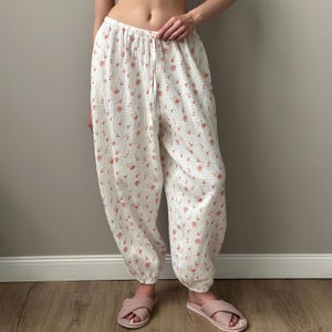 May include: White loose-fitting pants featuring a delicate pink floral pattern and a textured, crinkled fabric finish. The pants have an elasticized waistband with a matching drawstring tie and gathered cuffs at the ankles for a relaxed fit.