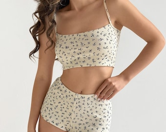 May include: A two-piece set consisting of a cropped tank top and high-waisted shorts. Both pieces are made from a cream-colored fabric featuring a small, repeating dark blue floral pattern. The top has thin shoulder straps and a straight neckline.