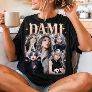 May include: A black short-sleeve t-shirt featuring a graphic print with the text DAMI in a stylized font. The design includes five photographic portraits of a person with various hair colors and poses, surrounded by a glowing, star-like light effect.