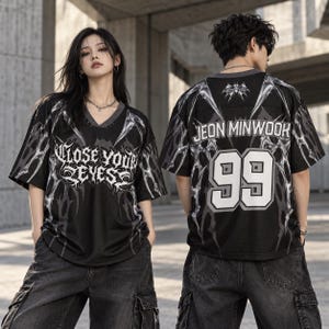 May include: A black V-neck jersey featuring a metallic silver web-like pattern, the text "CLOSE YOUR EYES" on the front, and the name "JEON MINWOOK" with the number "99" on the back. The shirt has short sleeves and a dark grey collar trim.