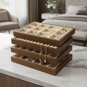 Può includere: Una pila di quattro vassoi rettangolari in legno per l'organizzazione di gioielli con finitura marrone scuro e rivestimento in velluto beige. Il vassoio superiore presenta diversi scomparti quadrati contenenti vari anelli e orecchini.