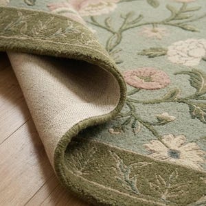 May include: A sage green area rug featuring a floral pattern with pink and white blossoms and green vines. The rug has a dark olive green border with a leaf motif. One corner of the rug is folded back, revealing a light beige woven backing material.