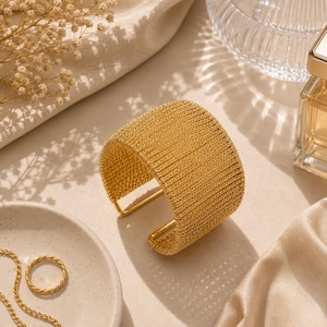 May include: A wide gold cuff bracelet featuring a textured, woven metal mesh design. The bracelet has an open-back construction with smooth, rounded edges. The surface of the metal has a bright, reflective finish that catches the light.