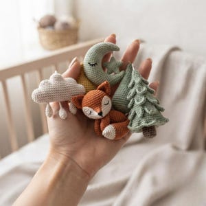 Könnte beinhalten: Eine Sammlung kleiner Häkelornamente, bestehend aus einer cremefarbenen Wolke mit Regentropfen, einer salbeigrünen Mondsichel mit Stern, einem rostorangen schlafenden Fuchs und einer salbeigrünen Tanne mit braunem Stamm, alles aus Garn.