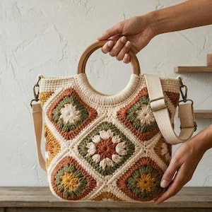 Puede incluir: Un bolso de ganchillo con un patrón de motivos cuadrados en tonos crema, verde oliva, amarillo mostaza y terracota. El bolso incluye un asa circular de madera y una correa de hombro ajustable de tela color crema con herrajes metálicos.