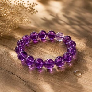 May include: A circular bracelet composed of smooth, polished, deep purple spherical beads. One bead is clear and colorless, contrasting with the uniform dark violet hue of the other beads. The bracelet is arranged in a ring shape on a wooden surface.