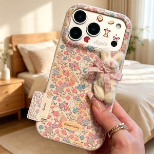 May include: A phone case featuring a floral pattern in shades of pink, blue, and yellow with small rabbit silhouettes. The case includes a plush white rabbit figure with a pink ribbon, a small tag labeled Rabbit, and a label reading Rabbit Garden.
