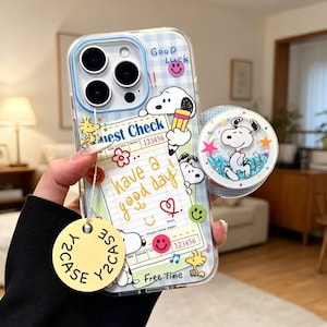 May include: A clear phone case featuring a blue plaid background, Snoopy and Woodstock illustrations, and a guest check graphic with the text 'have a good day'. It includes a circular liquid glitter pop-out grip and a round yellow Y2CASE tag.