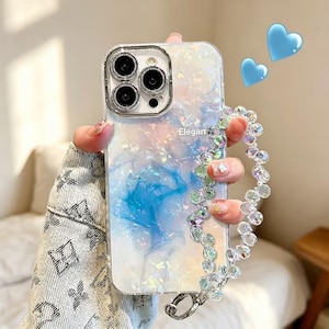 May include: A clear smartphone case with an iridescent, pearlescent finish featuring blue and white marbled patterns. The case includes the text "Elegant" and a detachable wrist strap made of faceted, clear, light-reflecting crystal-like beads.