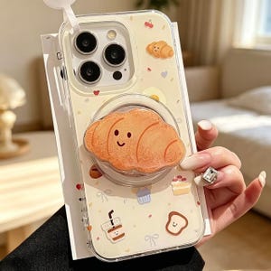 May include: A clear smartphone case featuring a light yellow background with small illustrations of cherries, croissants, cupcakes, and coffee cups. A large, circular, three-dimensional croissant-shaped grip is attached to the center back.