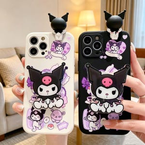 May include: Two smartphone cases, one white and one black, featuring a 3D Kuromi character attachment at the top and a larger Kuromi figure pop-out grip in the center. The cases are decorated with purple and white Kuromi character graphics.
