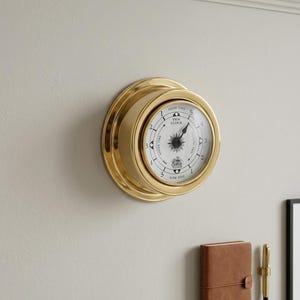 Puede incluir: Un reloj de mareas circular con un marco de latón pulido y una esfera blanca. La esfera presenta marcas negras, una rosa de los vientos central y el texto "TIDE CLOCK" con indicadores de "HIGH TIDE" y "LOW TIDE". Una manecilla negra señala la lectura.
