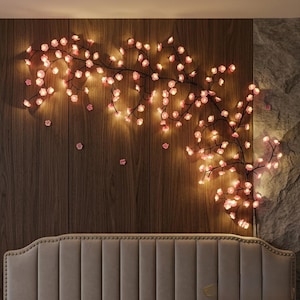Puede incluir: Una lámpara decorativa de pared con tallos oscuros y ramificados adornados con numerosas flores de cerezo rosas iluminadas. El arreglo floral brillante se extiende sobre una superficie de pared con paneles de madera.