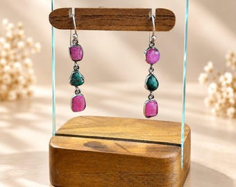 Può includere: Un paio di orecchini pendenti con tre pietre grezze ciascuno, sospesi a ganci in argento. Le pietre si alternano tra rosa vivace e verde intenso, tenute in posizione da montature in argento, esposti su un supporto in legno.
