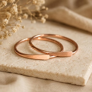 Puede incluir: Dos brazaletes finos de oro rosa pulido con una banda lisa y redondeada y una sección final ligeramente aplanada y cónica. Las pulseras presentan un acabado metálico cálido y un diseño circular simple y continuo que se superpone ligeramente.