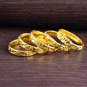 Può includere: Un set di quattro anelli d'oro disposti in fila, ognuno caratterizzato da un motivo in rilievo continuo di piccoli fiori e volute. Gli anelli hanno una finitura metallica lucida e sono posizionati leggermente di sbieco per mostrare i dettagli.
