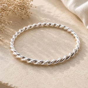 Può includere: Un bracciale rigido in argento lucido caratterizzato da un design continuo a corda intrecciata. Il cerchio metallico presenta una superficie riflettente con solchi profondi che creano una trama a spirale lungo l'intera circonferenza.