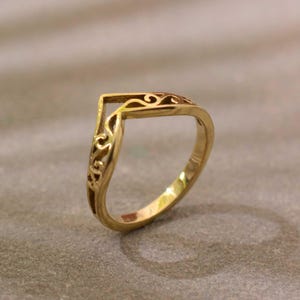 May include: A gold-coloured V-shaped ring featuring an openwork filigree design along the band. The ring has a pointed chevron silhouette with intricate cut-out scrollwork patterns visible on the sides and top surface of the metal band.