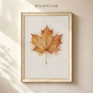 Pode incluir: Uma impressão de arte emoldurada com uma ilustração em aguarela de uma única folha de ácer castanha centrada num fundo creme claro. O texto PEACEFUL OAK PRINTS está impresso a preto no topo central do passe-partout branco sujo.