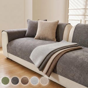 Könnte beinhalten: Ein helles Sofa, das mit einem dunkelgrauen, strukturierten Stoffschutz bedeckt ist. Das Set enthält ein passendes quadratisches Kissen in einem hellen Beigeton. Fünf kleine kreisförmige Farbmuster sind unten im Bild zu sehen.