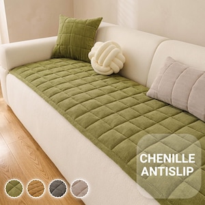 Puede incluir: Una funda de sofá rectangular de chenilla verde oliva con un patrón de rejilla acolchado y reverso antideslizante. La funda está colocada sobre un sofá color crema, acompañada de un cojín cuadrado a juego, un cojín de nudo crema y uno beige.