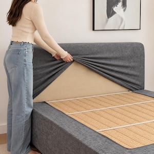 Puede incluir: Una funda de tela gris oscuro para cabecero se coloca sobre un cabecero beige. La funda presenta una apariencia tejida y texturizada, diseñada para estirarse sobre la estructura existente. El marco de la cama es visible con una esterilla.