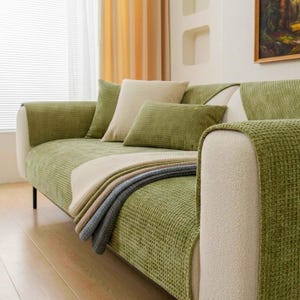 May include: A sofa covered in a textured, olive green waffle-knit fabric. The couch features a cream-coloured throw pillow, a matching olive green rectangular pillow, and a layered throw blanket in shades of cream, beige, and grey.