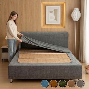 Puede incluir: Una funda de tela gris oscuro para estructura de cama con acabado texturizado, que incluye un cabecero rectangular y una base que envuelve el marco. La funda se muestra parcialmente levantada para revelar los listones de madera.