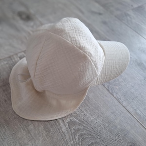 Può includere: Un cappello da sole color crema con una finitura in tessuto testurizzato e stropicciato. Il cappello presenta una calotta arrotondata, una tesa anteriore corta e una falda posteriore estesa progettata per coprire la zona del collo. Un cordino è visibile.