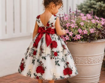 May include: A sleeveless white dress featuring an embroidered pattern of red roses and green stems, a deep V-shaped back, and a large dark red satin bow at the waist. The dress is paired with simple white flat shoes with a single strap.